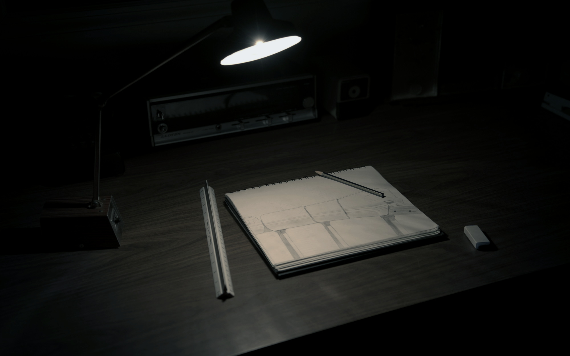 Un carnet ouvert sur un tracé technique, éclairé par une lampe articulée ; un réglet et un crayon posés dessus, le reste de la pièce dans le noir.